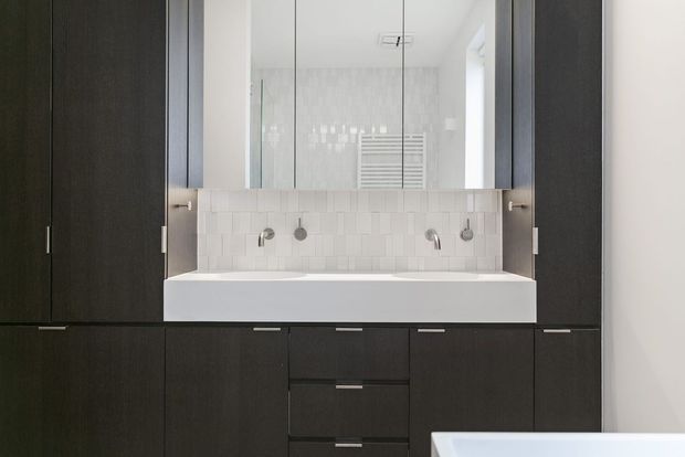 Contemporary bathroom vanity with a wide white integrated basin set into dark timber cabinetry. A white rectangular tile splashback, wall-mounted chrome fixtures, and a large mirror create a clean, high-contrast composition. The edge of a white bathtub is visible in the foreground.