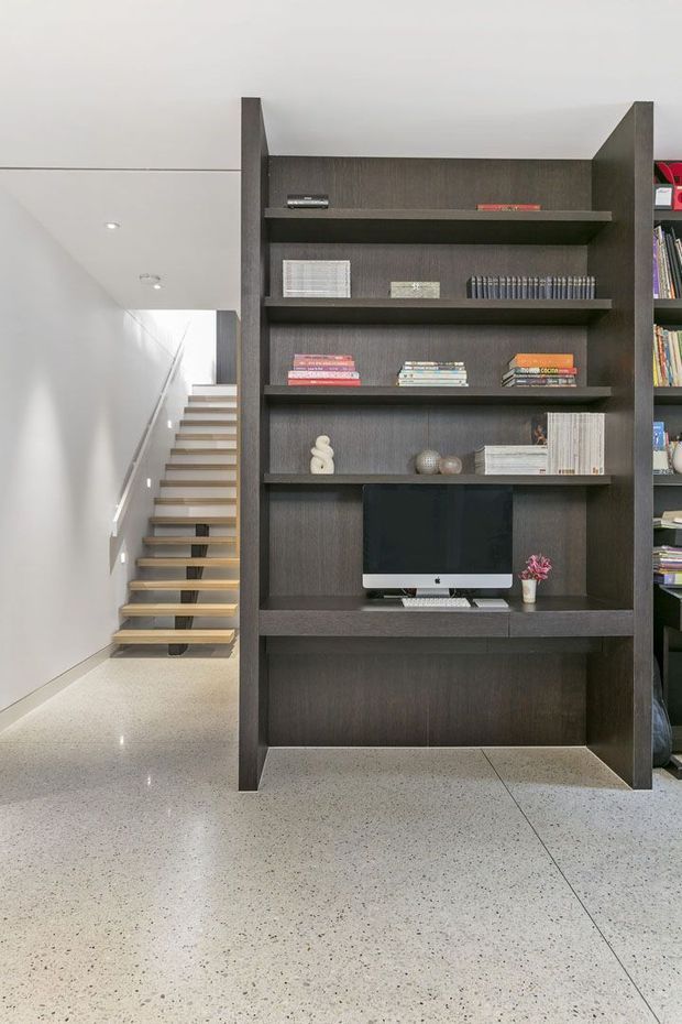 Contemporary interior view featuring a tall dark timber built-in shelving and media unit, with books, decorative objects and a computer display. A minimalist open staircase with timber treads and a glass or slim balustrade rises behind it, set against white walls and a polished light terrazzo floor.