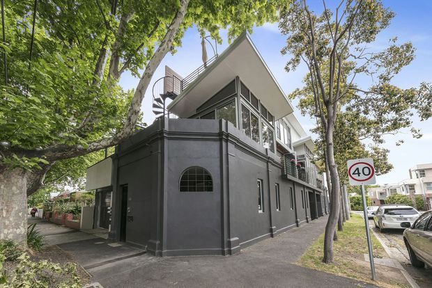 Street-level view of a contemporary multi-storey dark-rendered building with a distinctive angular white roof, extensive upper-level glazing, balconies, and a curved metal stair enclosure. The building sits on a leafy urban suburban street with mature trees and adjacent residential properties.