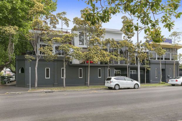 Street-level exterior view of a contemporary multi-storey residential or commercial building with a dark grey lower facade, light upper volumes, extensive windows, balconies, and mature street trees. Parked cars and an urban streetscape are visible in front.