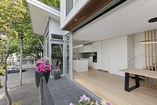 Modern open-plan kitchen and dining area viewed from a paved outdoor terrace through fully opened glazed doors. The interior features white flat-panel cabinetry, integrated appliances, pale timber flooring, a light timber dining table with black legs, minimalist linear pendant lighting, and vertical timber screening. Large aluminium-framed glazing and a projecting roof structure connect the interior to the leafy urban exterior.