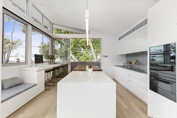 Bright contemporary white kitchen with a large central island, integrated appliances, white cabinetry, pale timber flooring, and extensive floor-to-ceiling glazing. A built-in window seat and dining area are visible beside the kitchen, with leafy outdoor views beyond.