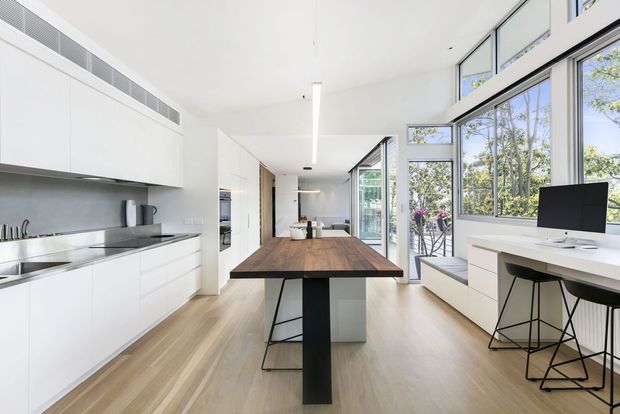 Bright open-plan contemporary interior combining a streamlined white kitchen with integrated appliances, a large timber dining island, and a compact home office along expansive black-framed windows. Pale timber flooring, white cabinetry, generous glazing, and views to greenery create an airy, minimal living space.