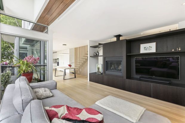 Bright contemporary open-plan living room with a large grey sectional sofa, pale timber flooring, and a dark timber built-in media wall incorporating a fireplace and television. Floor-to-ceiling glazing opens to a leafy outdoor setting, while a timber-lined ceiling section and vertical timber screen add warmth and texture.
