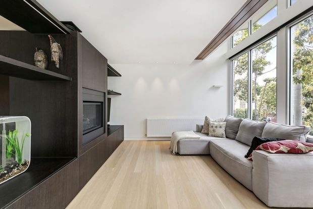 Bright contemporary living room with a large grey sectional sofa, pale timber flooring, a dark built-in entertainment and display wall, white walls, and expansive floor-to-ceiling windows overlooking trees. A slim timber-lined ceiling feature adds warmth and definition.