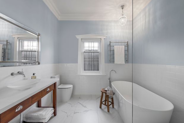 Bright, elegant bathroom with pale blue-grey walls, white subway-tile wainscoting, marble-look tiled flooring, a freestanding white bathtub, pedestal-style basin on a timber vanity, toilet, towel ladder, and leaded-glass window. A large mirror enhances the sense of space, while chrome fixtures and a crystal pendant add refined traditional detailing.