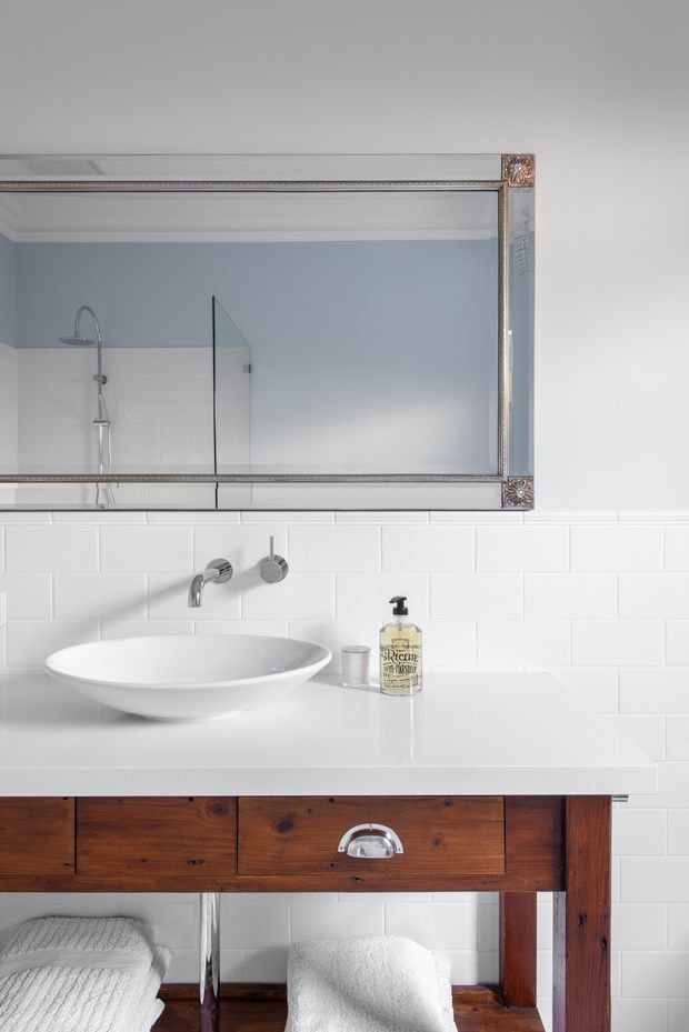 A bright, refined bathroom vanity featuring a white vessel basin on a polished white benchtop, wall-mounted chrome tapware, white subway tile backsplash, and a rustic dark timber open vanity with drawers. A large ornate framed mirror reflects a frameless glass shower enclosure and pale blue bathroom wall. White towels and a soap dispenser complete the scene.