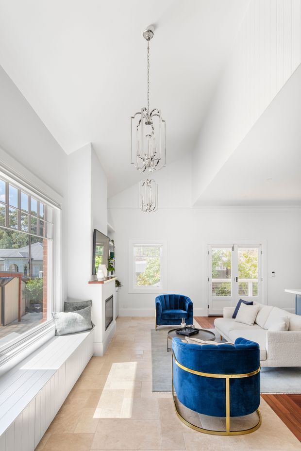 Bright, refined living room with a high angled white ceiling, tall multi-pane windows, white built-in window seating, pale stone-look flooring, and a contemporary fireplace surround. A white sofa and vivid cobalt-blue velvet lounge chairs with brass trim frame a central coffee table. Crystal-style pendant lights hang prominently from the ceiling, and timber flooring is visible at the edge of the room.