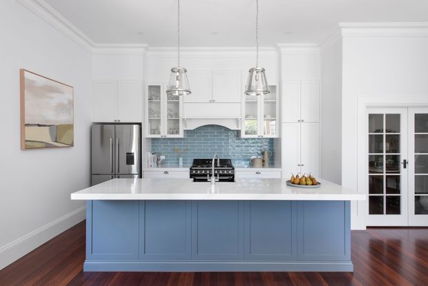 Bright, refined kitchen with a large blue-painted island, white perimeter cabinetry, a pale engineered-stone benchtop, stainless-steel appliances, and a blue-grey subway tile splashback. Clear pendant lights hang above the island, with dark timber flooring, high decorative ceiling cornices, and French-style glazed doors visible at right.