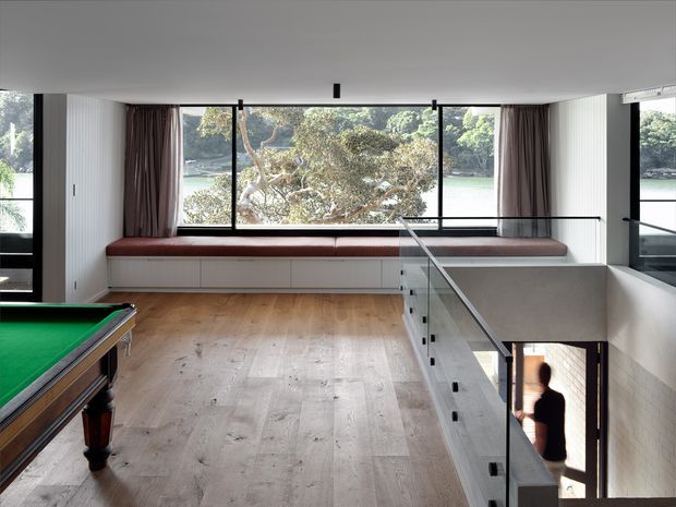 A bright contemporary upper-level living or recreation space with a timber floor, panoramic black-framed windows overlooking greenery and water, and a long built-in window seat upholstered in deep burgundy. A pool table occupies the foreground, while a glass balustrade borders an open stairwell leading to the level below. Vertical white wall panelling and dark curtains add contrast to the restrained interior.
