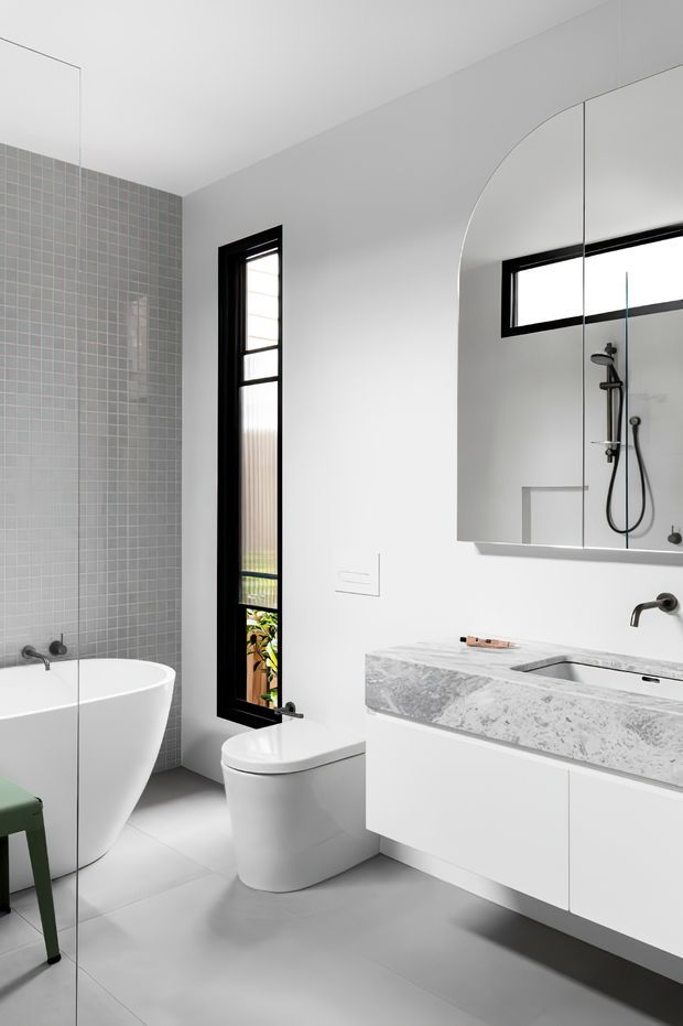 A bright contemporary bathroom featuring a freestanding white bathtub, wall-mounted white vanity with a veined stone benchtop, integrated basin, black tapware, a frameless arched mirror, a wall-hung toilet, and a glass-enclosed shower visible in the reflection. The room has pale grey mosaic wall tiles, smooth light grey floor tiles, white walls, and black-framed vertical windows.