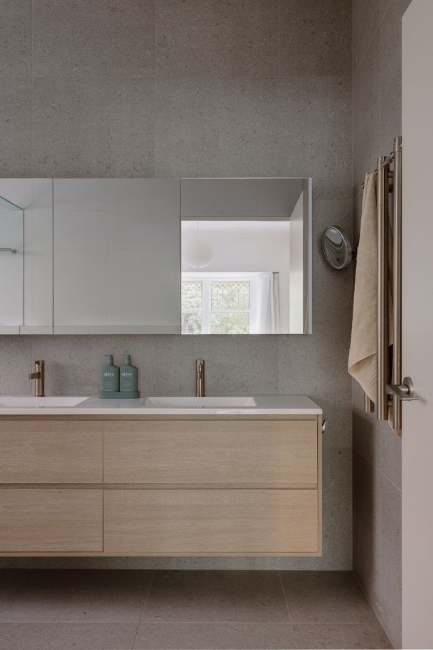 Contemporary bathroom vanity with a wide floating light-wood cabinet, white integrated double basins, brushed brass tapware, and a large mirrored wall cabinet. Large-format grey stone-look tiles cover the walls and floor. A heated towel rail with beige towels is mounted beside the doorway, while a window and pendant light are visible in the mirror reflection.