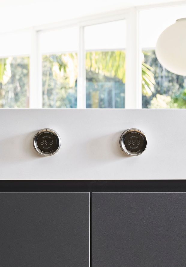 Close-up interior detail of a sleek modern kitchen or cooking appliance installation. A white horizontal panel contains two round digital controls with metallic rims, above dark charcoal textured cabinetry. Large white-framed windows provide bright natural light, and a spherical pendant light is partially visible at upper right.