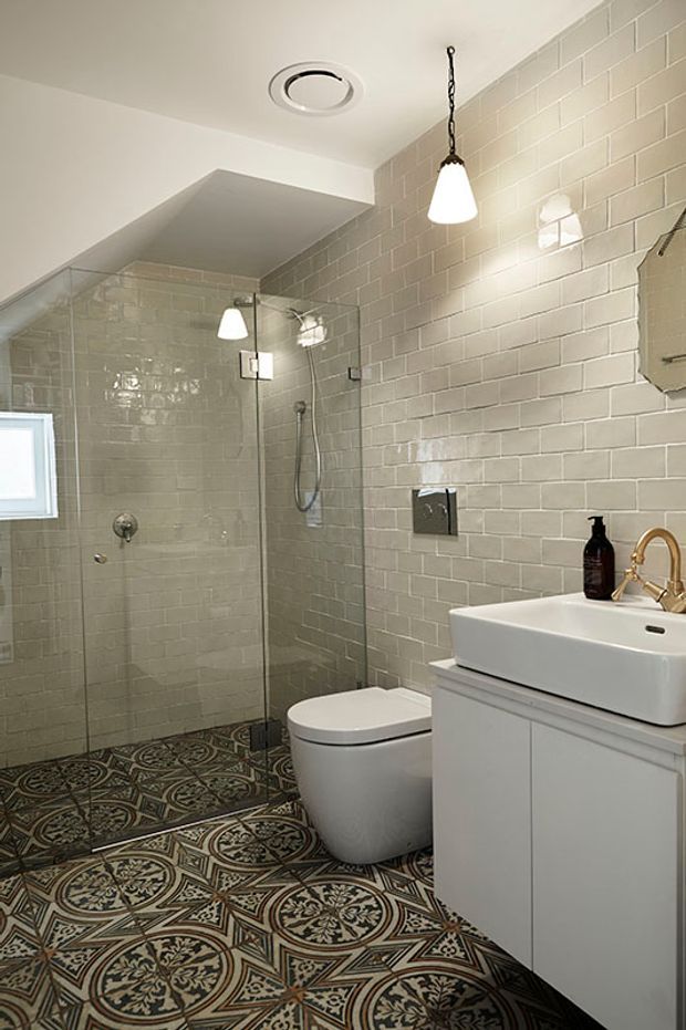 A compact, bright bathroom featuring a walk-in glass shower with handheld showerhead, wall-hung toilet, white vessel basin on a white vanity, brass tapware, pale grey glossy subway-tile walls, and patterned encaustic-style floor tiles. A hanging pendant and recessed ceiling vent provide lighting and ventilation.