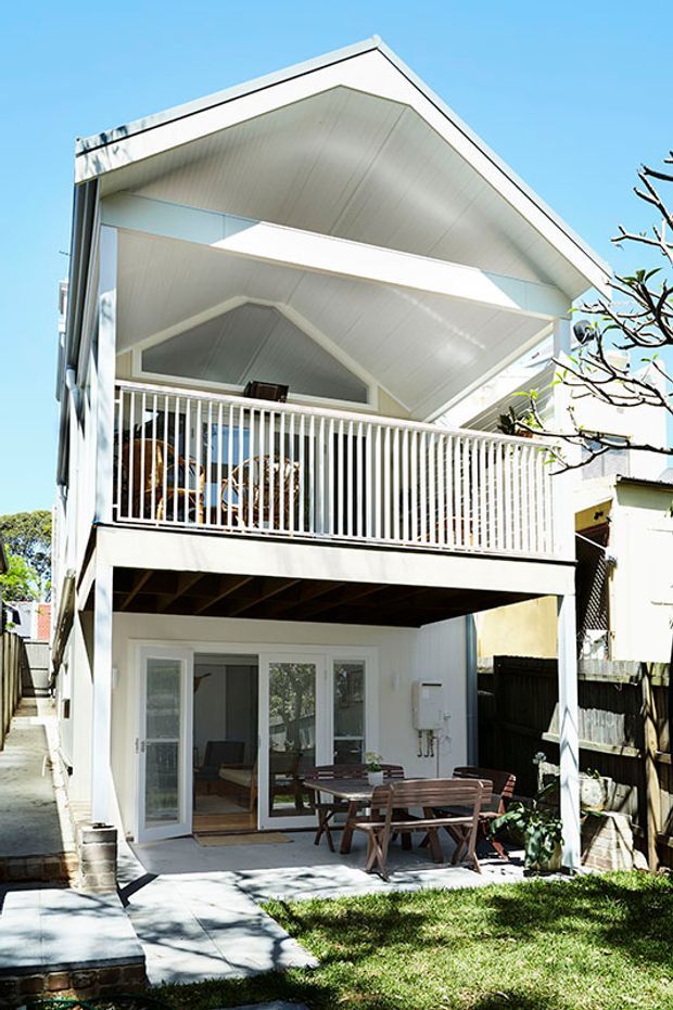 Exterior view of a narrow, multi-level white house with a prominent steep gable roof and deep covered upper balcony. White vertical balcony railings, pale cladding, exposed lower-level structure, glazed doors, and a ground-level outdoor dining area are visible. The design has a bright contemporary coastal character and appears suited to a compact urban or suburban site.