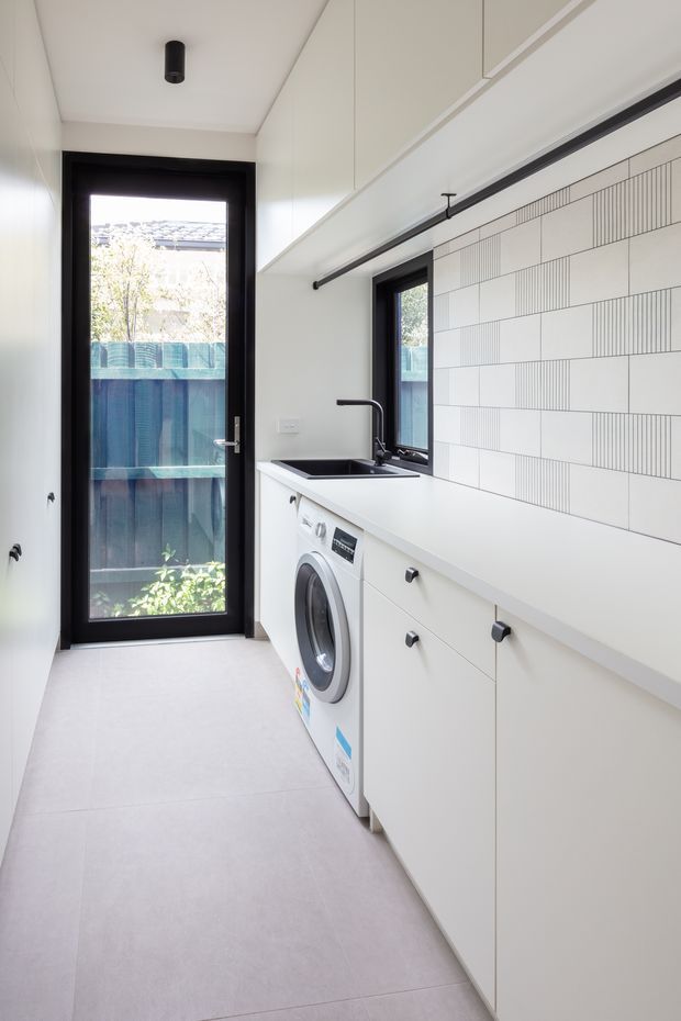Bright, narrow contemporary laundry room with full-height white cabinetry, a white benchtop, integrated front-loading washing machine, black undermount sink and tap, and a linear patterned white tile splashback. Black-framed glazed door and window provide natural light, while pale large-format floor tiles and black hardware create a crisp monochrome scheme.