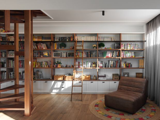 A warm, light-filled library or reading room with extensive built-in timber shelving spanning the wall, filled with books and display objects. A rolling ladder provides access to upper shelves, while a low built-in cabinet runs beneath them. The room has timber flooring, a brown leather lounge chair, a colourful circular rug, dark full-height curtains, and a partially visible timber stair structure at left. The design blends contemporary detailing with mid-century-inspired timber joinery.