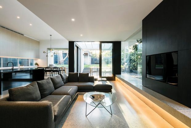 A refined open-plan living space with a large charcoal sectional sofa and glass coffee table in the foreground, opening to a dining area and sleek kitchen. Floor-to-ceiling glazing and black-framed doors connect the room to an outdoor courtyard, while a dark built-in media wall with fireplace anchors the right side. Warm timber flooring, pale walls, recessed lighting, and contemporary detailing create a bright, polished interior.