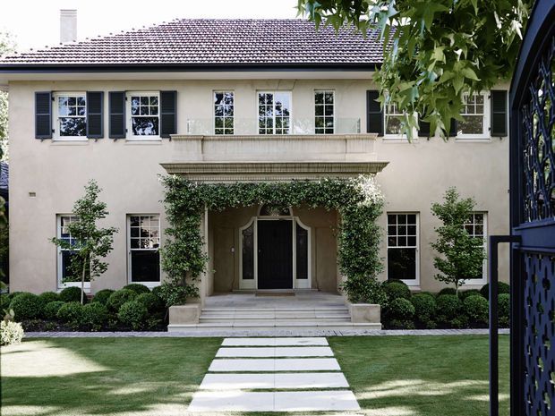 Symmetrical two-storey classical-style residential facade viewed from the front garden. The rendered cream facade features a tiled hipped roof, multi-pane windows with dark louvred shutters, a formal central entry with a projecting balcony and decorative cornice, climbing greenery, clipped hedges, lawn, and a pale stepping-stone path. A dark ornamental gate frames the right edge.