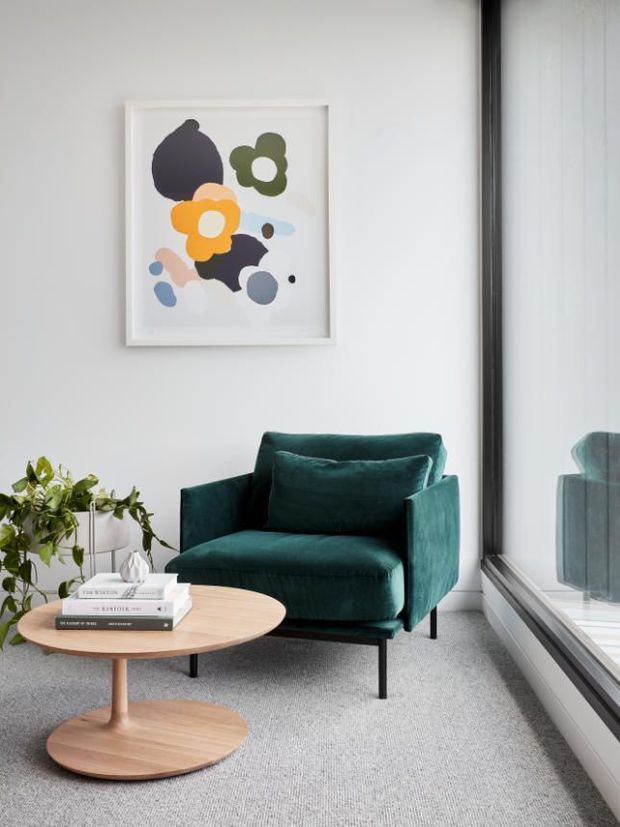 A bright, minimalist sitting area beside full-height glazing, featuring a deep teal upholstered armchair, round light-wood pedestal coffee table, grey carpet, white walls, a potted trailing plant, and colourful abstract artwork. The large window reflects the chair and fills the space with natural light.