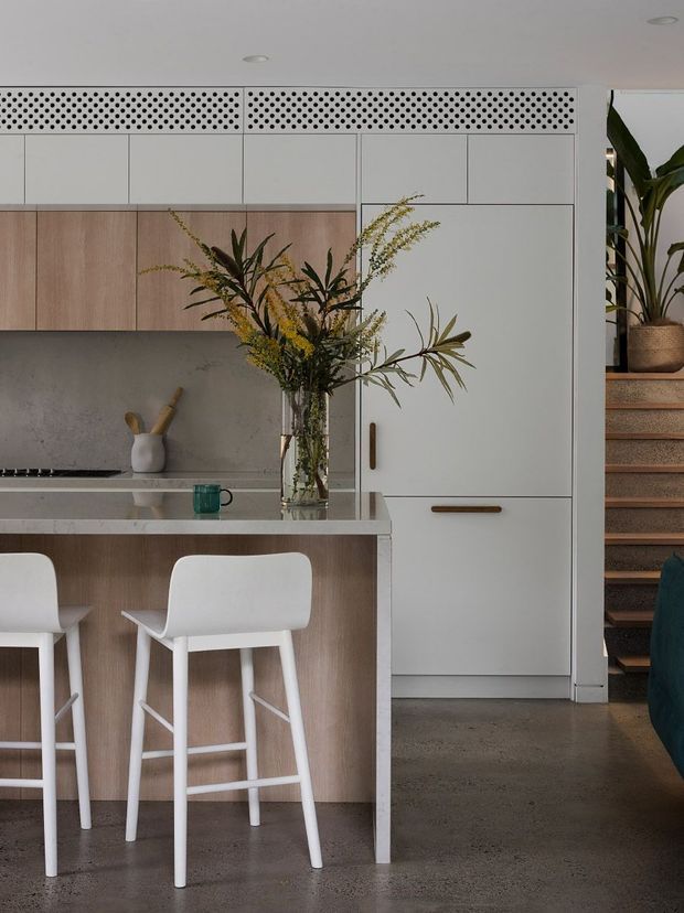 A refined contemporary kitchen with a compact island and white bar stools. The kitchen features pale timber cabinetry, white upper cabinets, a grey concrete-look splashback, integrated tall storage, perforated ventilation panels above the cabinetry, and a polished concrete floor. A staircase and large indoor plant are visible at the right edge.