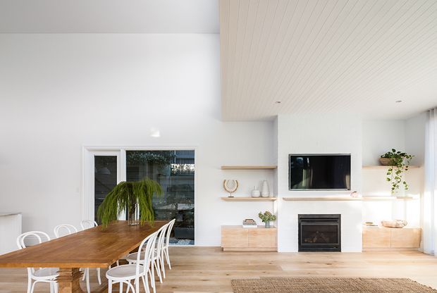 Bright open-plan living and dining area with white walls, pale timber flooring, a long timber dining table, white dining chairs, built-in timber cabinetry, open shelving, a wall-mounted television and a fireplace. The room has a clean contemporary feel, with a vertically panelled timber ceiling feature and abundant natural light.