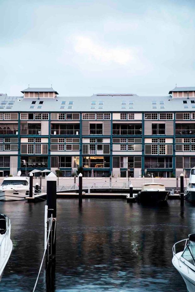 Exterior view across a marina toward a multi-storey waterfront building with a pale blue-grey roof, repetitive gridded windows, balconies, louvred panels, and teal structural accents. Boats, docks, and dark reflective water occupy the foreground.