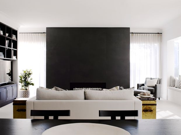 Bright contemporary living room viewed from behind a large light-colored sofa, with a dramatic dark feature wall and fireplace, built-in shelving and cabinetry at left, sheer curtains, armchairs, and a low dark coffee table. The room has strong black-and-white contrast with warm metallic accent side tables.