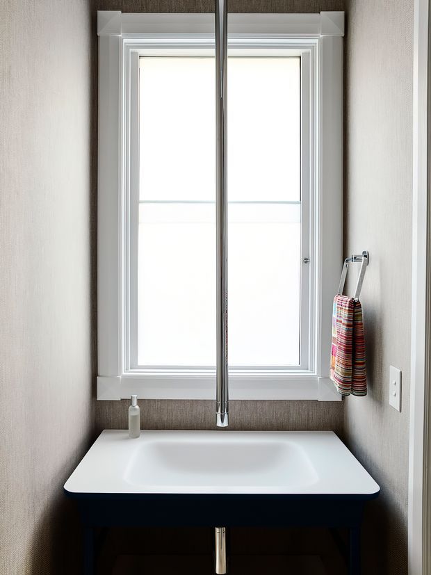Compact, bright bathroom washbasin area with a white sculptural basin, dark vanity base, tall central mirror, white-trimmed window, textured taupe wall finish, and a colorful striped hand towel.