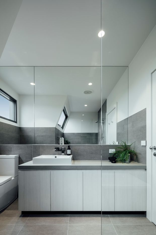 Bright, contemporary bathroom viewed through a frameless glass shower screen. The room features large grey wall and floor tiles, a floating light-wood vanity with a dark grey benchtop, a white vessel basin, wall-mounted black tapware, mirrored cabinetry or wall mirror, recessed ceiling lights, a toilet, and a small indoor plant. A high window and skylight-like ventilation elements provide additional light and ventilation.