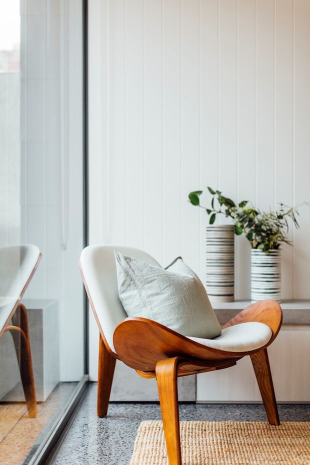 A bright, refined interior corner featuring a mid-century-style timber lounge chair with white upholstery and a pale cushion beside floor-to-ceiling glazing. White vertical wall panelling, terrazzo flooring, a low built-in ledge with striped ceramic planters, and a natural woven rug create a calm, warm setting.