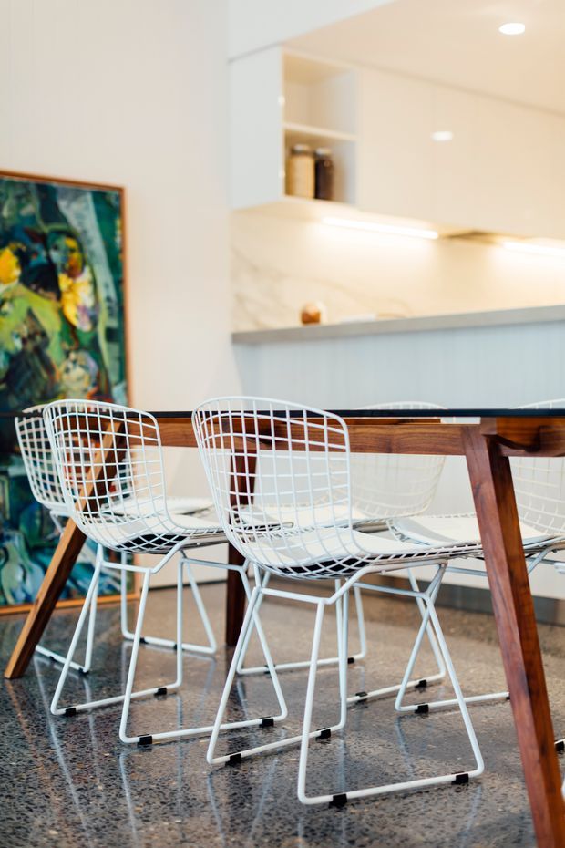 A bright open-plan dining area with a mid-century-style timber dining table and white wire-frame chairs. The space opens toward a contemporary kitchen with pale cabinetry, a light stone or marble-look splashback, under-cabinet lighting, and open shelving. A colourful framed artwork leans against the wall, while the polished speckled floor reflects the furniture and light.