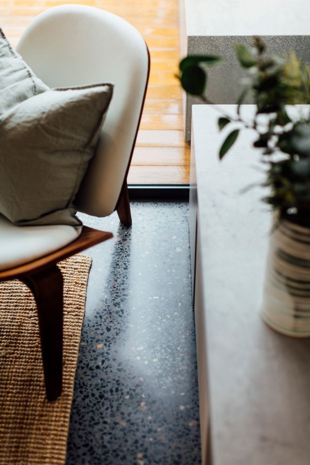 Close-up interior detail of a refined living area, showing a cream upholstered lounge chair with a neutral cushion, dark timber and woven cane framing, a terrazzo floor, and a pale stone or concrete-topped console with a potted plant. Warm timber flooring and a large window or glazed opening provide natural light.