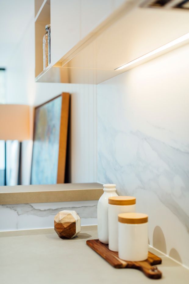 Close-up interior view of a refined bathroom vanity area with a pale marble-look wall finish and matching splashback, a large mirror, integrated linear under-cabinet lighting, and a light neutral benchtop. Ceramic containers and a wooden tray sit on the vanity, while a framed artwork and lamp are softly reflected in the mirror.