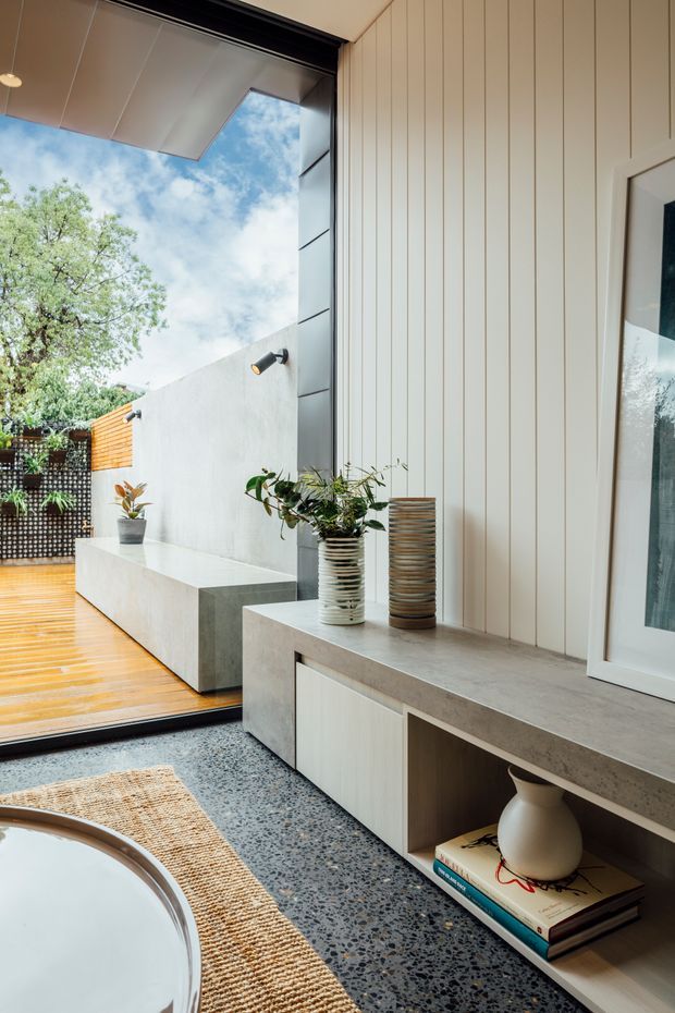 A refined bathroom interior opening onto a sunny outdoor courtyard. The room features vertical white wall panelling, a terrazzo floor, a long concrete-look built-in vanity or console with pale cabinetry, decorative pottery, and a partially visible freestanding bathtub. Large dark-framed glazing connects the bathroom to a timber-decked courtyard with a concrete bench and planted screen. The overall aesthetic is calm, contemporary, and natural.