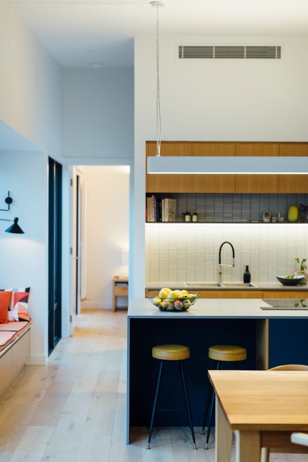 Bright contemporary kitchen viewed from an adjacent open-plan area, featuring a dark navy island with pale stone benchtop, timber cabinetry and shelving, a white small-format tiled splashback, integrated linear pendant lighting, black tapware, and two stools. A timber dining table and a colourful built-in window seat are visible at the edges, with a hallway extending beyond the kitchen.