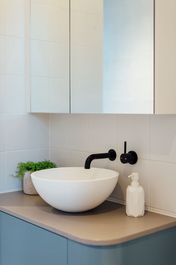 Close-up of a contemporary bathroom vanity featuring a white round vessel basin on a taupe countertop, matte black wall-mounted tapware, pale blue cabinetry, white tiled walls, and simple white overhead cabinetry. A small plant and soap dispenser add restrained styling.