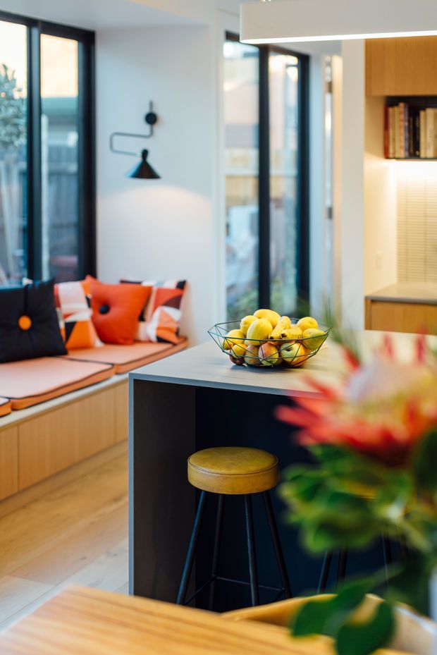 A bright, contemporary open-plan living and kitchen space with a compact dark island or breakfast counter in the foreground. The room features a built-in window seat with colourful geometric cushions, large black-framed windows, pale timber flooring, warm timber cabinetry, open shelving, and a pendant light. A fruit bowl and bar stool sit on the island, with indoor plants adding greenery.