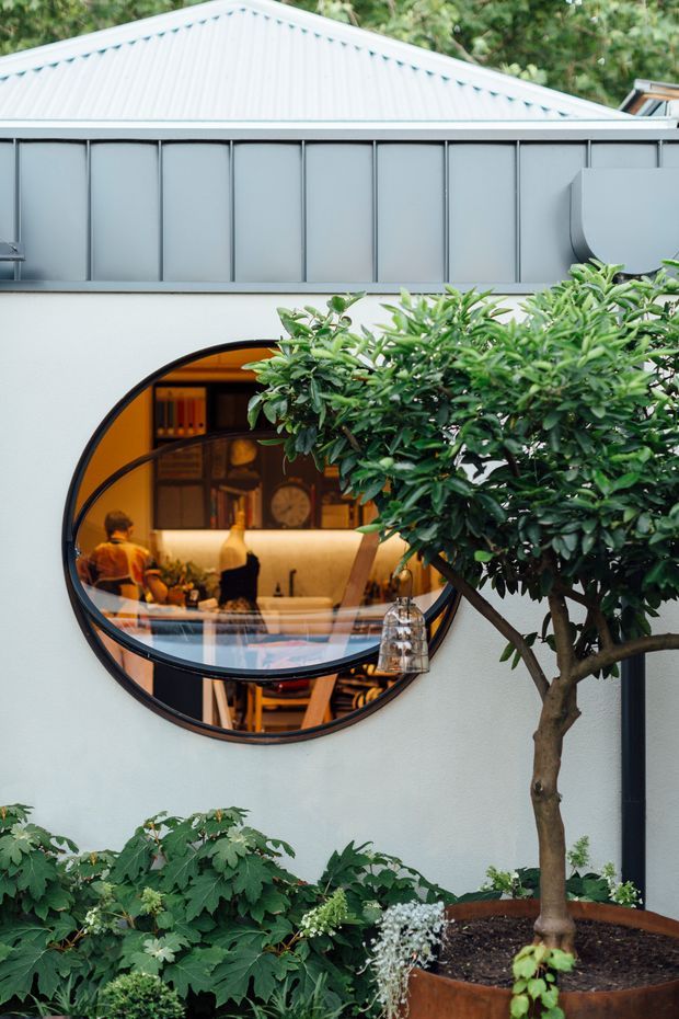 Exterior view of a contemporary building facade with pale rendered walls, grey standing-seam metal cladding, and a distinctive large circular window. The window reveals a warmly lit interior kitchen or dining area, while lush planted greenery and a potted tree occupy the foreground.