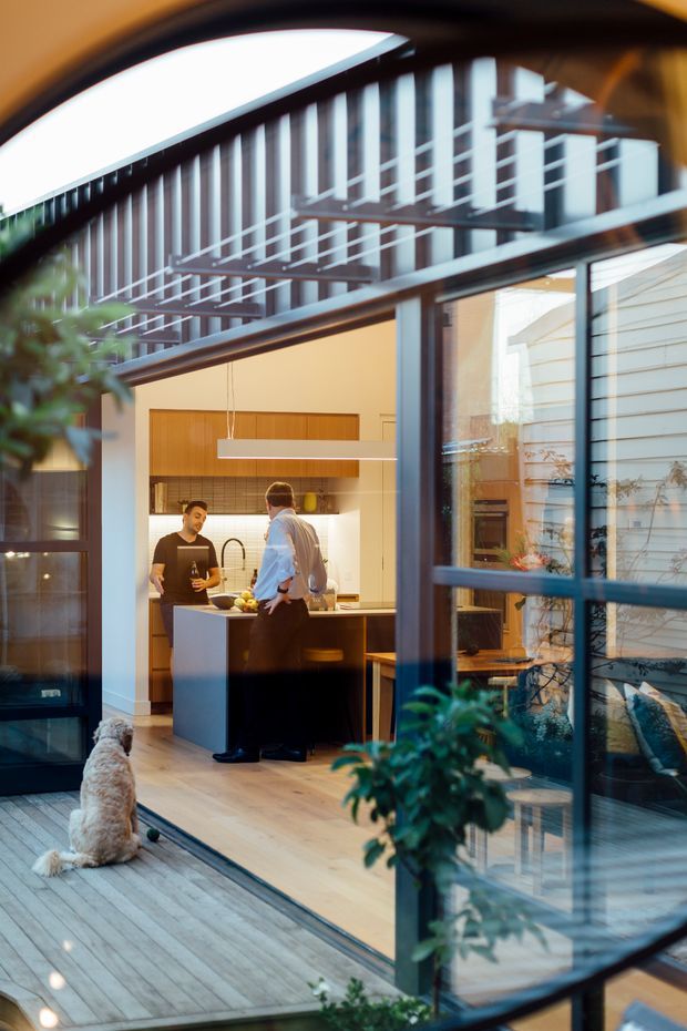 A warm, contemporary kitchen viewed from a timber deck through expansive black-framed glazing. Two people stand at a central island, with timber cabinetry, a pale benchtop, pendant lighting and integrated appliances visible inside. The image also shows a small courtyard-like outdoor space, a seated dog, lush potted greenery, and strong reflections of the surrounding structure in the glass.
