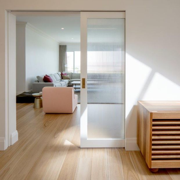 Bright interior view through a wide doorway into a contemporary living room. A white-framed sliding door with translucent ribbed glass separates the spaces, while pale timber flooring continues throughout. The living area contains a light sectional sofa, colourful cushions, a soft pink armchair and a low coffee table. In the foreground, a timber slatted cabinet sits against a white wall, with strong geometric sunlight and shadow adding contrast.