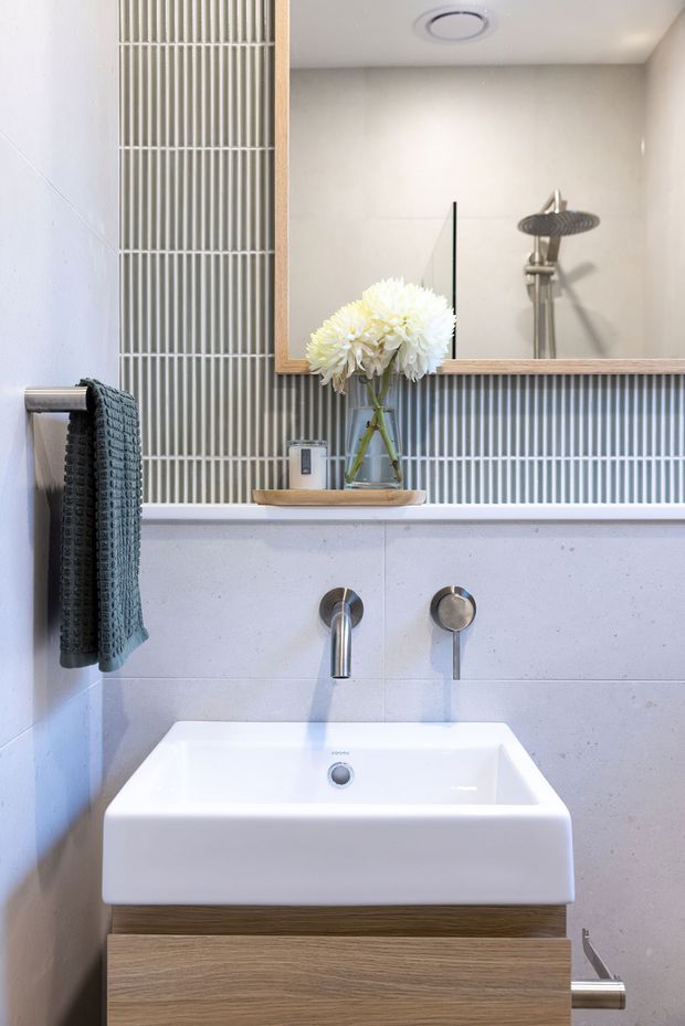 Compact contemporary bathroom vanity with a white rectangular basin, wall-mounted chrome tapware, pale stone-look wall tiles, vertically ribbed grey feature tiles, and a large timber-framed mirror reflecting the shower. A timber vanity cabinet and muted teal towel add warmth and contrast.
