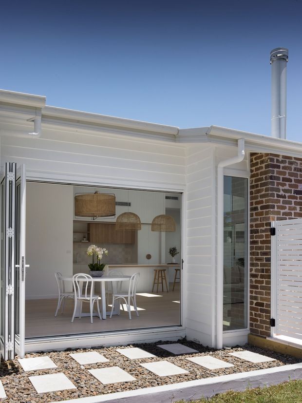 Bright exterior view of a contemporary single-storey home extension with white horizontal weatherboard cladding, a low-pitched roof, white-framed bi-fold glazing, and an exposed brick side wall. The open doors reveal a dining area and kitchen with timber cabinetry and woven pendant lights. A gravel courtyard with large rectangular stepping stones sits in the foreground, and a metal flue projects above the roof.