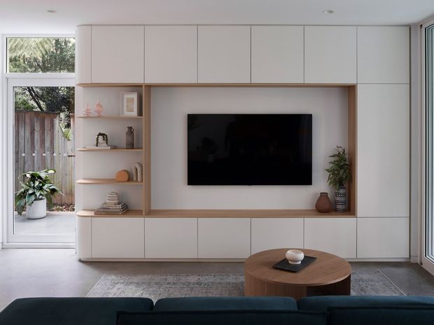 Contemporary living room with a built-in full-height media wall and integrated storage cabinetry in a soft neutral finish. The joinery features a recessed timber-toned TV niche, open display shelves, and a wall-mounted television. A dark sofa, round timber coffee table, patterned rug, indoor plants, and large glazed doors/windows create a calm, refined space with indoor-outdoor connection.