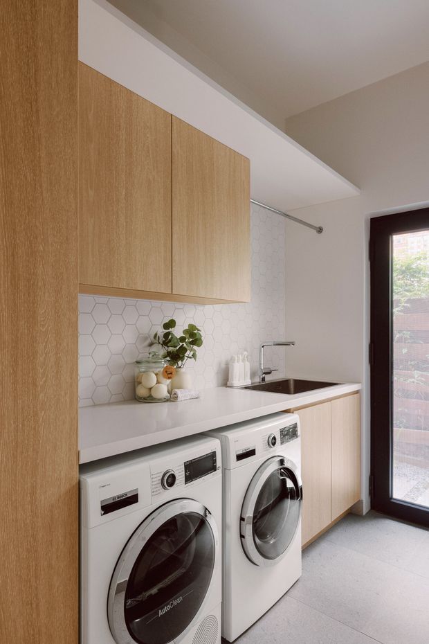 A compact, contemporary laundry room with warm light-wood cabinetry, a white engineered-stone countertop, white hexagonal tile splashback, integrated sink and tap, and front-loading white washer and dryer. A dark-framed glazed exterior door provides natural light. The room has a clean, practical, built-in design with pale grey flooring and neutral walls.