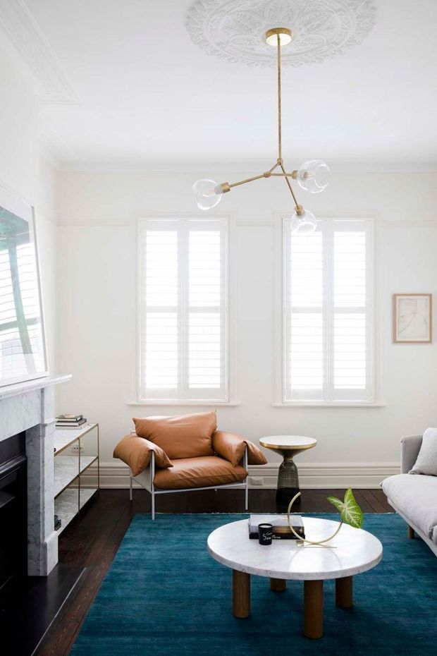 Bright, refined living room with white decorative wall and ceiling detailing, tall plantation-shuttered windows, dark timber flooring, and a marble fireplace surround. A cognac leather armchair, pale upholstered sofa, round marble coffee table, turquoise rug, brass-and-glass pendant, and small side table create a contemporary interior with traditional architectural character.