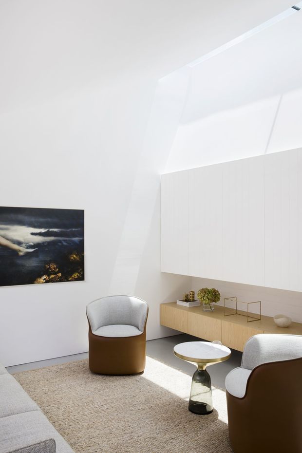 Bright, minimalist living room with a sculptural white angular ceiling and a high clerestory-like opening. The room features white walls, a long pale timber built-in console beneath white vertical-panel cabinetry, rounded brown-and-white lounge chairs, a pale sectional sofa, a textured neutral rug, and a small round side table. A dark landscape artwork provides contrast on the wall.