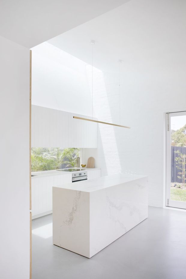 Bright minimalist white kitchen with a large waterfall-edge island in pale veined stone, flat-panel white cabinetry, integrated appliances, and a slim linear pendant light. High white ceilings and expansive glazing create an airy, clean space with views to greenery outside.