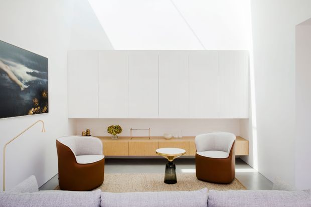 Bright, minimalist living room with a long built-in timber sideboard beneath full-height white cabinetry. Two rounded brown-and-cream lounge chairs face a small sculptural coffee table, with a pale sectional sofa in the foreground, textured rug, and a wall-mounted artwork. The room has a clean contemporary fit-out and a high, angled ceiling.