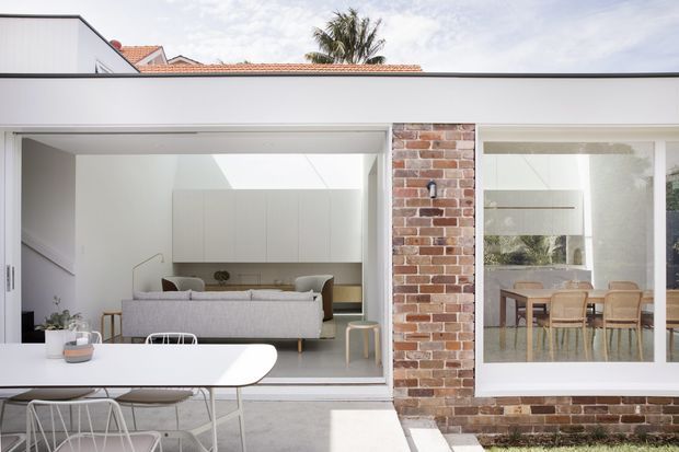 Bright contemporary rear facade with large glazed openings connecting an outdoor dining terrace to an open-plan living and dining interior. The white rendered structure is interrupted by a warm exposed brick pier, with a low flat roofline and glimpses of a red tiled roof and palm beyond. A grey sofa, pale built-in cabinetry, dining furniture, and white outdoor table and chairs are visible.